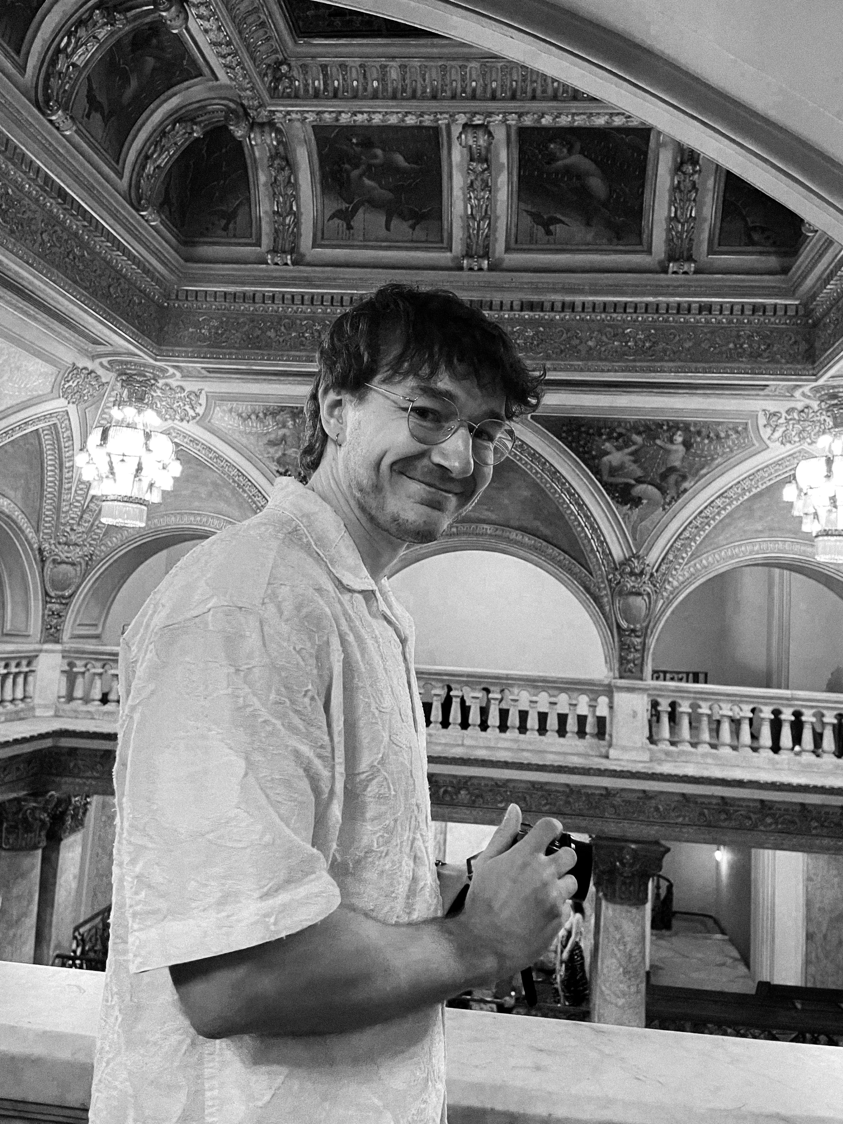 Black-and-white portrait of Jan Lauer at the balustrade of a historic building, camera in hand, with painted ceiling frescoes and chandeliers in the background.