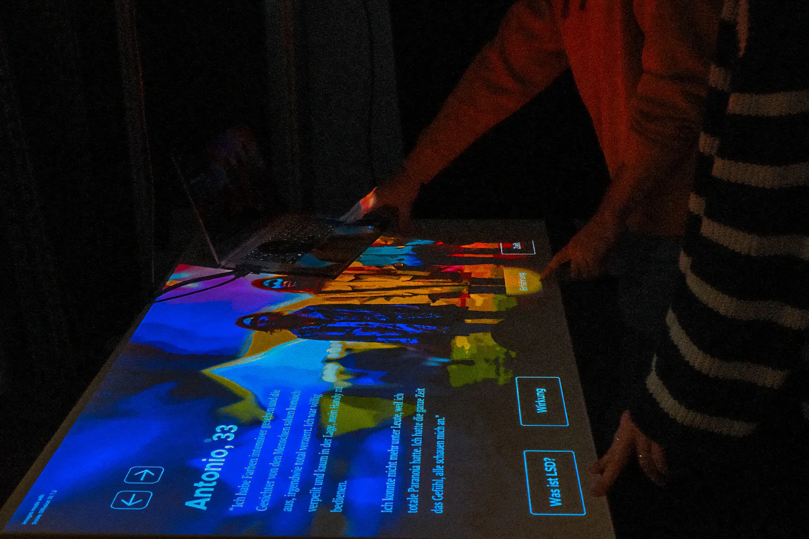 The projected exhibition table during testing: a hand touches the glowing touchpoints while a laptop sits on the tabletop.