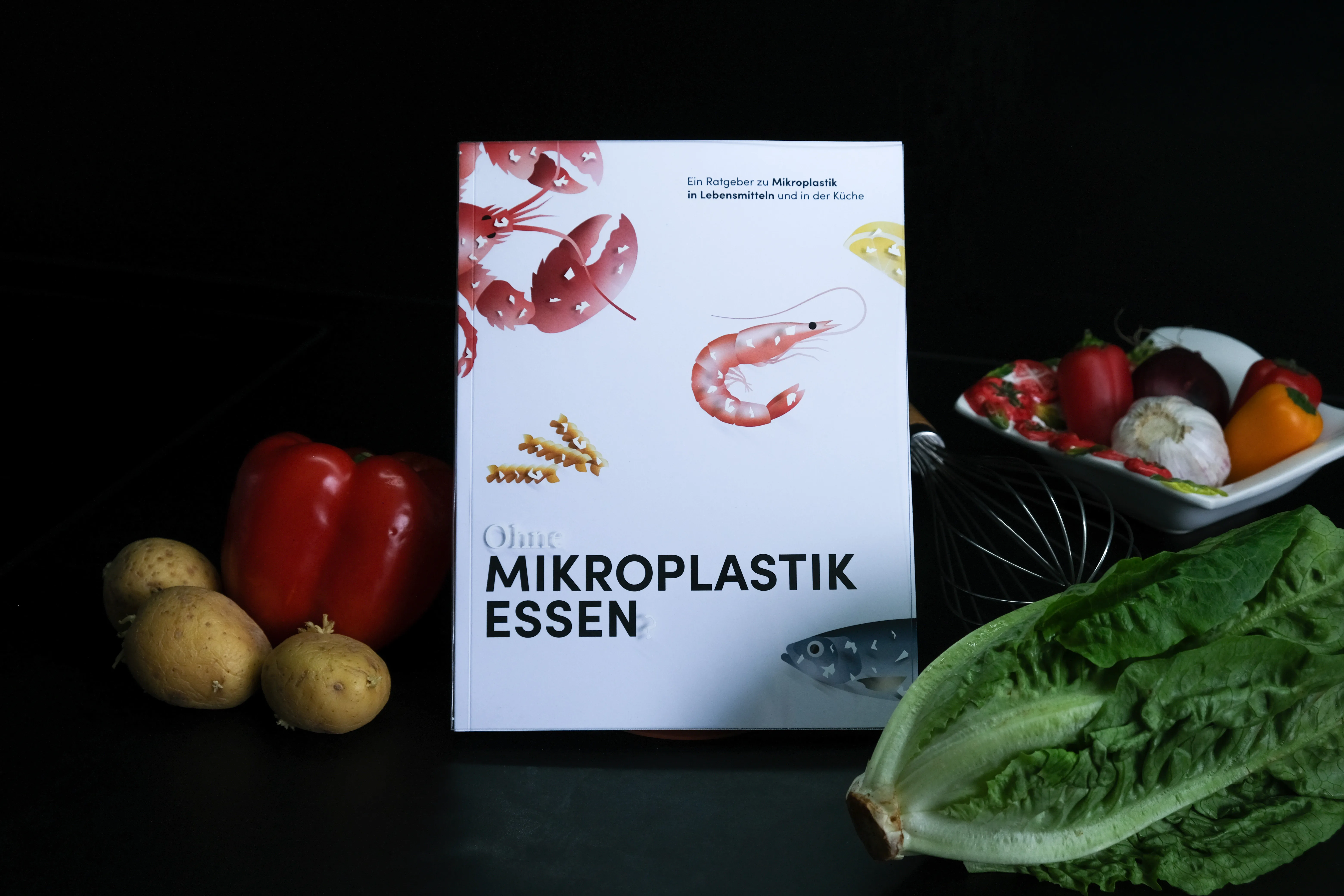 Photograph of the book cover in a dark kitchen setting, surrounded by vegetables and kitchen utensils - the cover features seafood illustrations with printed white plastic particles.